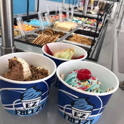 Three cups of ice cream with toppings in front of a dessert buffet, with various toppings and treats behind.