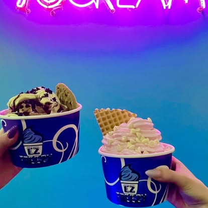 Two hands holding cups of ice cream with colorful toppings against a blue background with pink neon sign.