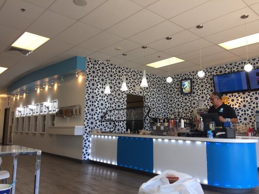 Coffee shop counter with a barista preparing drinks, decorated with patterned wall and illuminated ceiling lights.