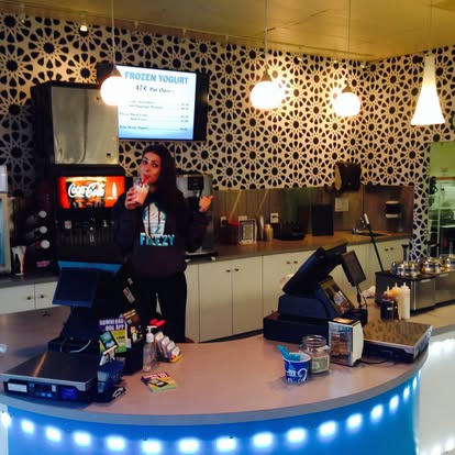 Person standing behind a curved counter with a blue LED light, in a cafe or fast-food setting, with a patterned wall and menu screen behind.