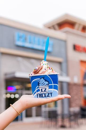 Hand holding a cup of ice cream with a blue spoon outside a shopping center, blue and white cup with EZ Freezy logo.