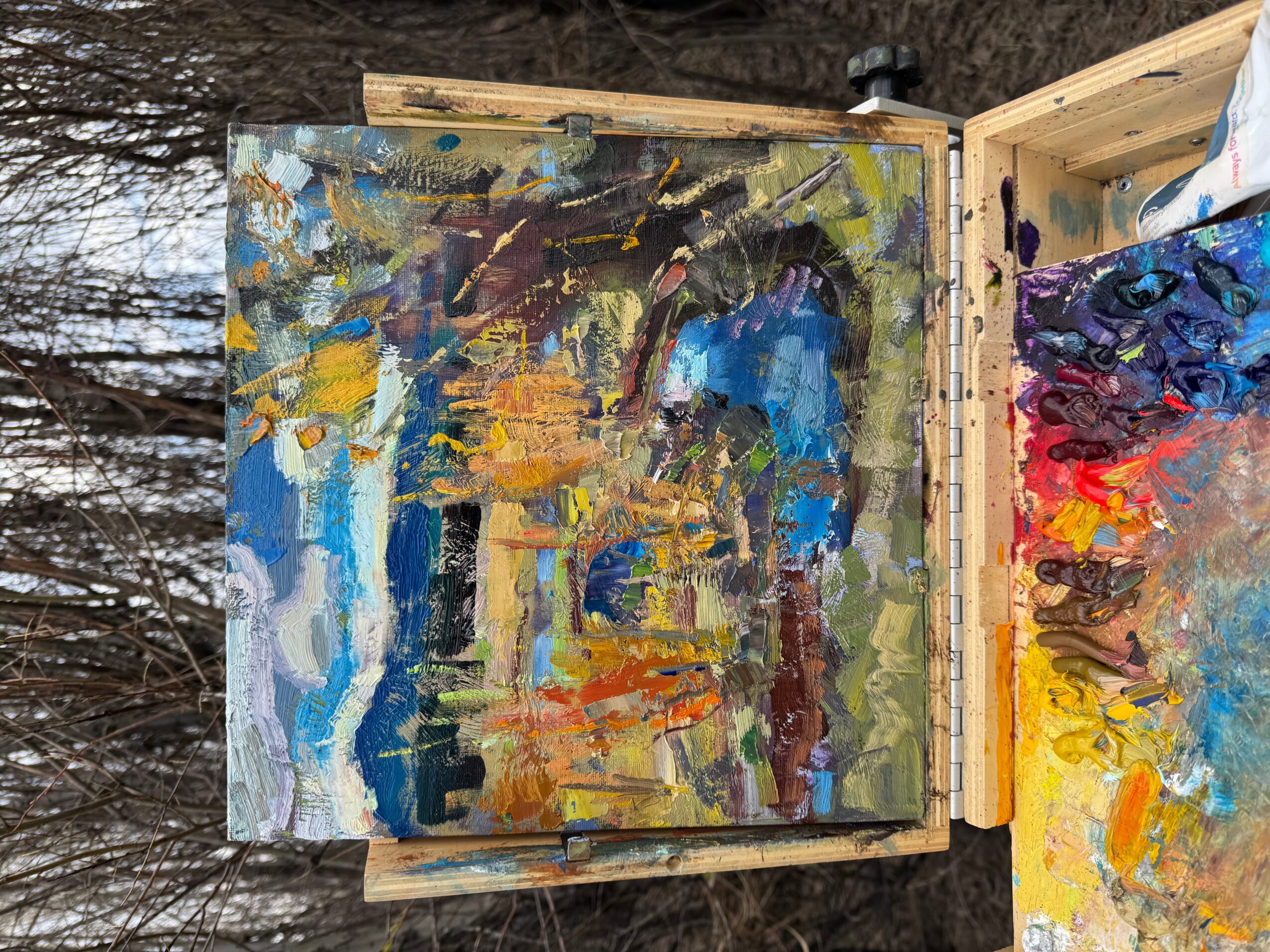 Abstract painting with blue, yellow, brown, and green colors on canvas, outdoors with trees in background.