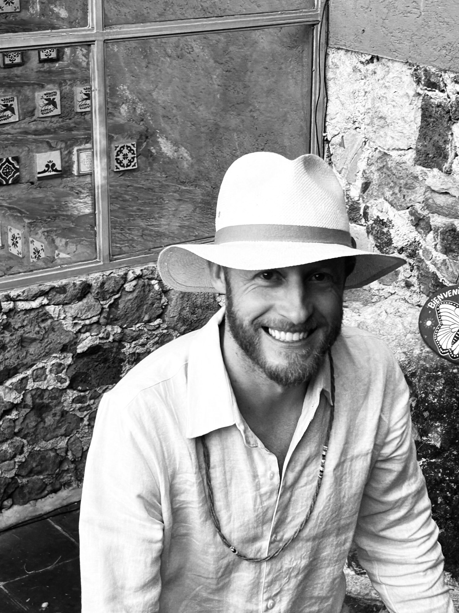 Smiling man wearing a hat and light-colored shirt, sitting outdoors with stone and brick background.