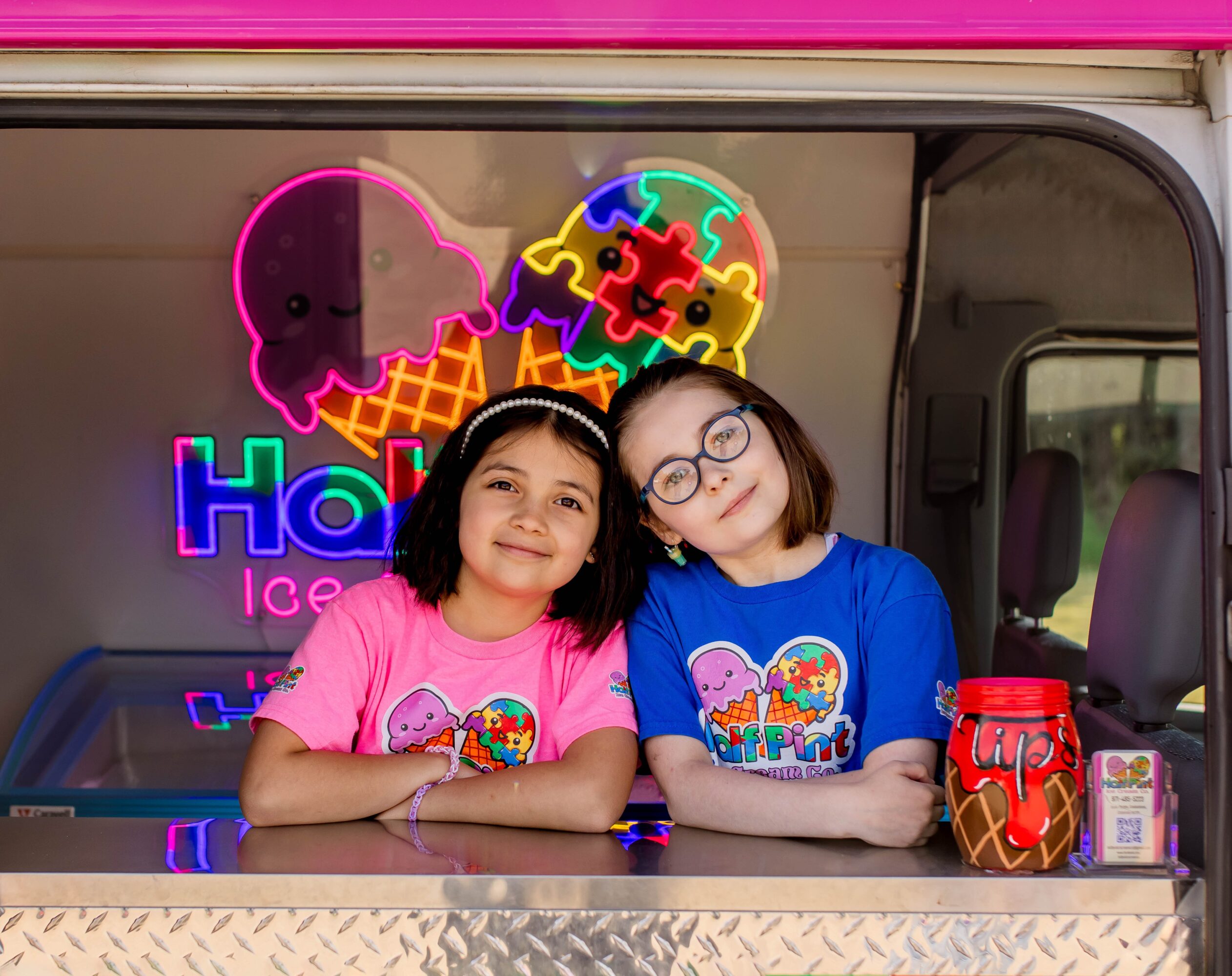 Two children with ice cream shirts leaning on a counter in front of a neon ice cream sign.