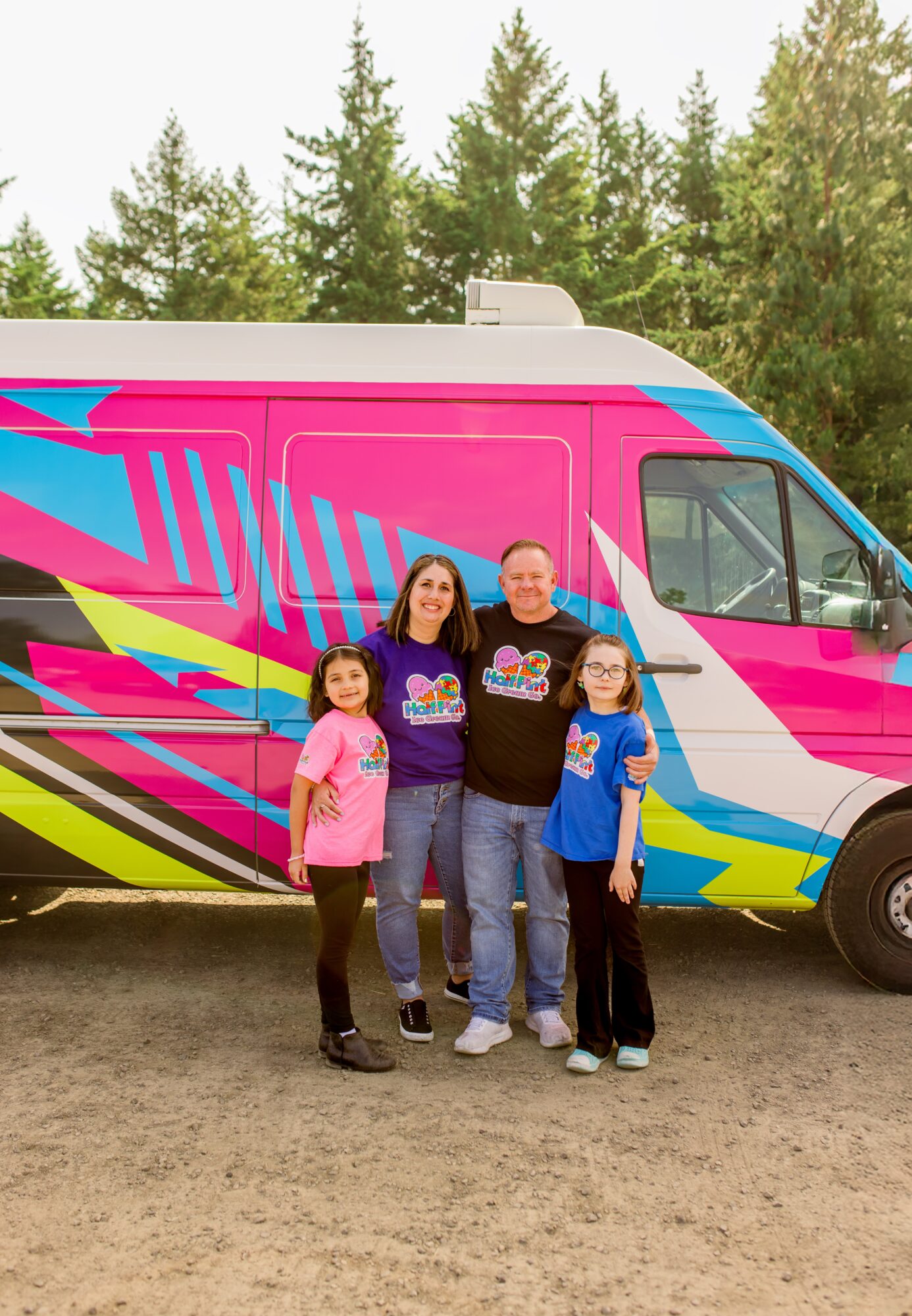 Four people stand in front of a colorful van with trees in the background, smiling at the camera.