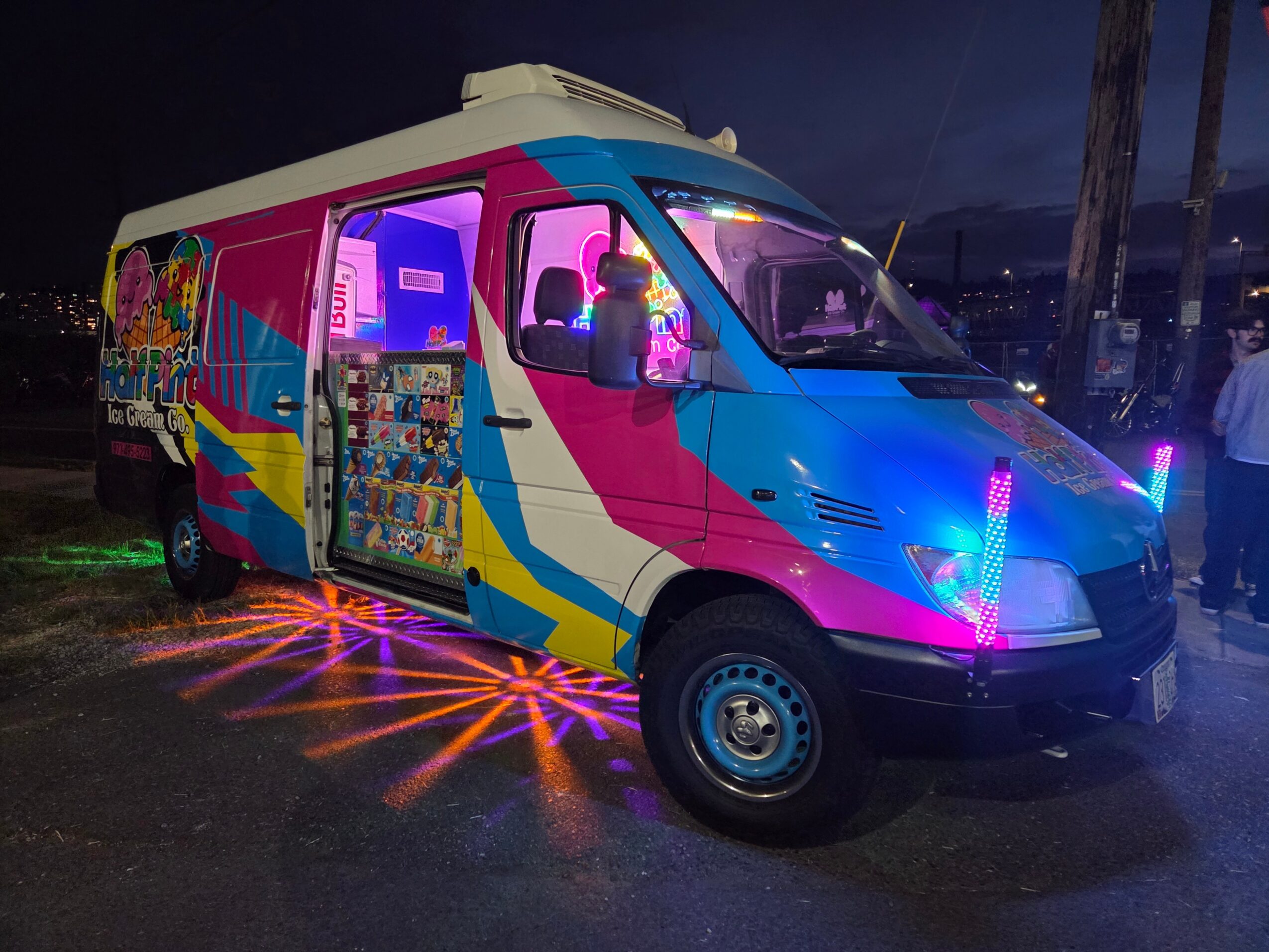 Colorful van with bright neon lights and artwork, parked outdoors at night, illuminated by vibrant lighting effects.