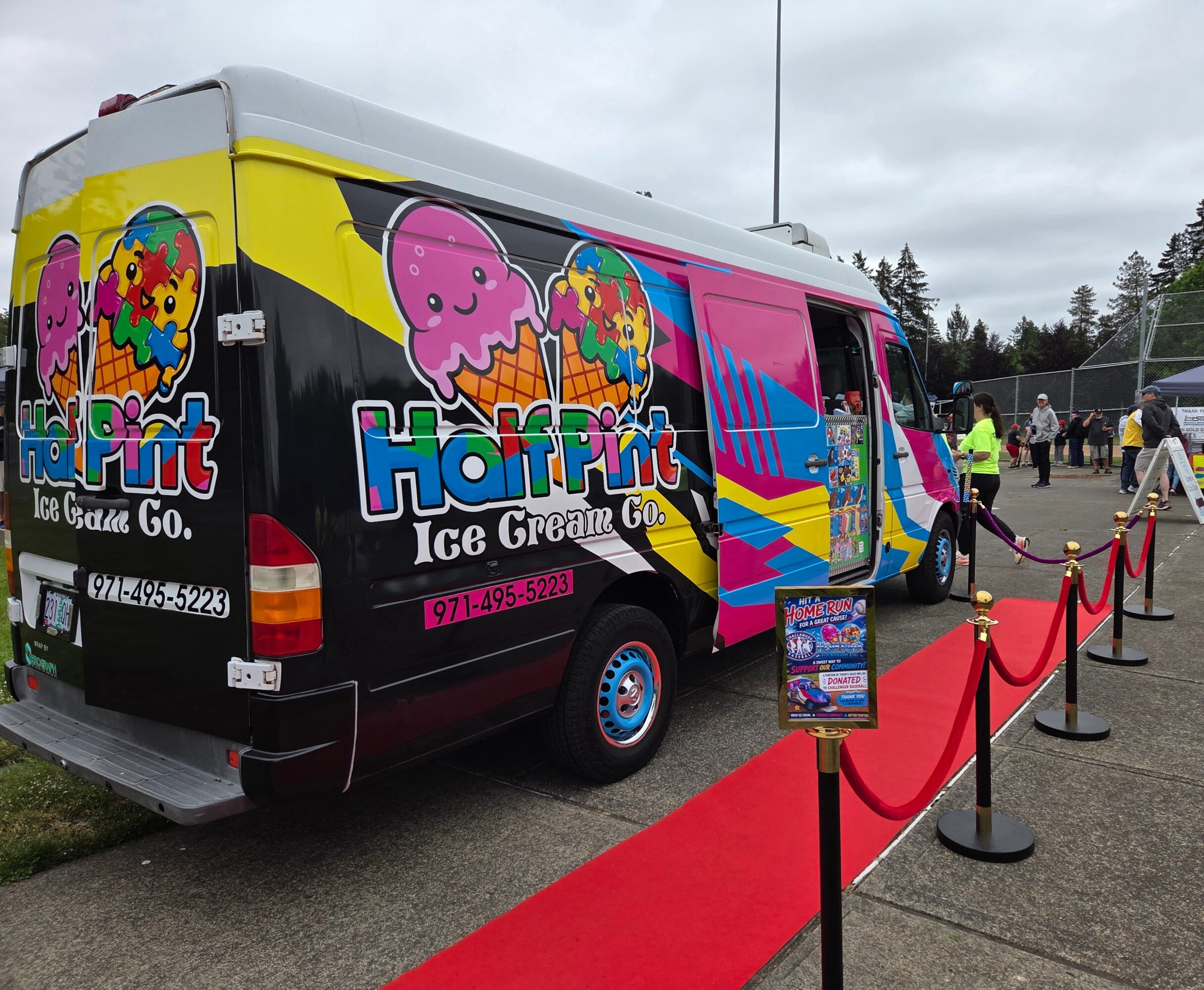 Ice cream truck with colorful graphics and logo, parked on a red carpet with people and trees in background.