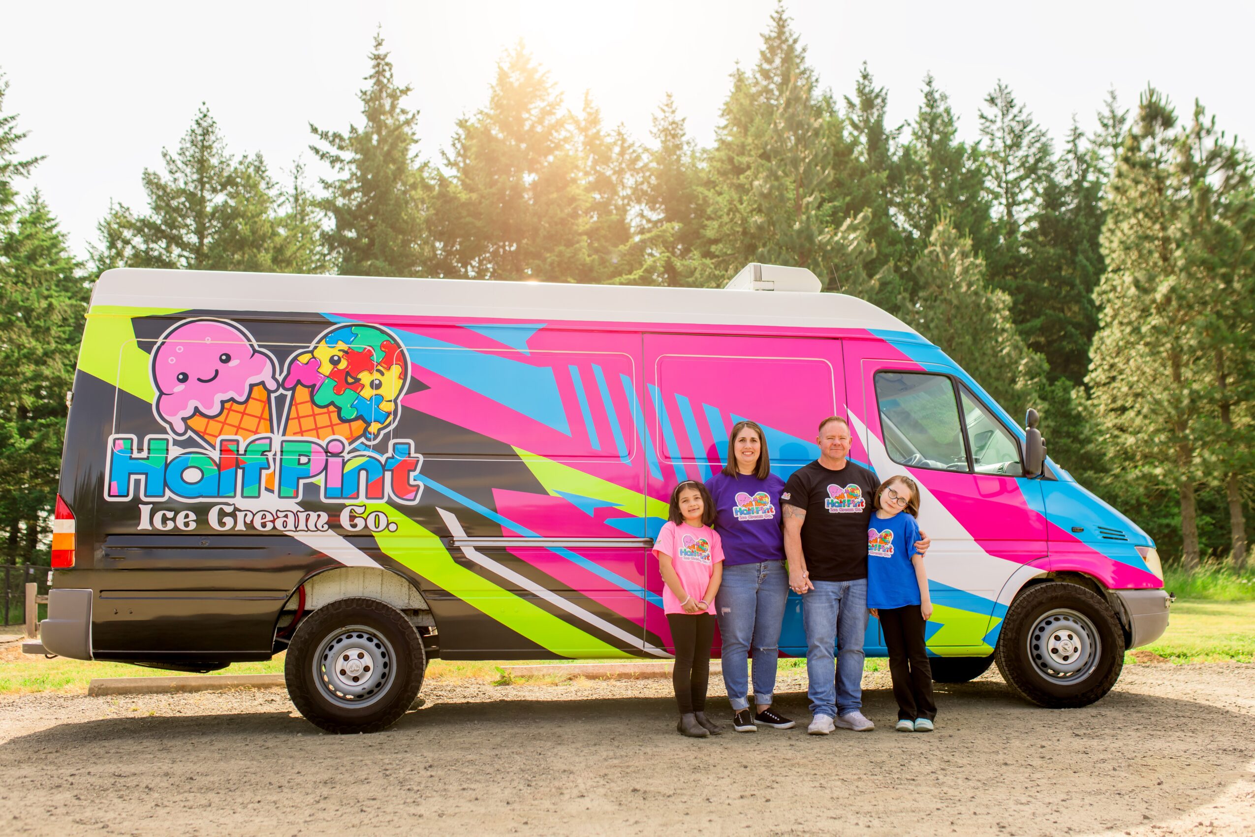 Four people stand in front of a colorful ice cream truck with trees and sky in the background.