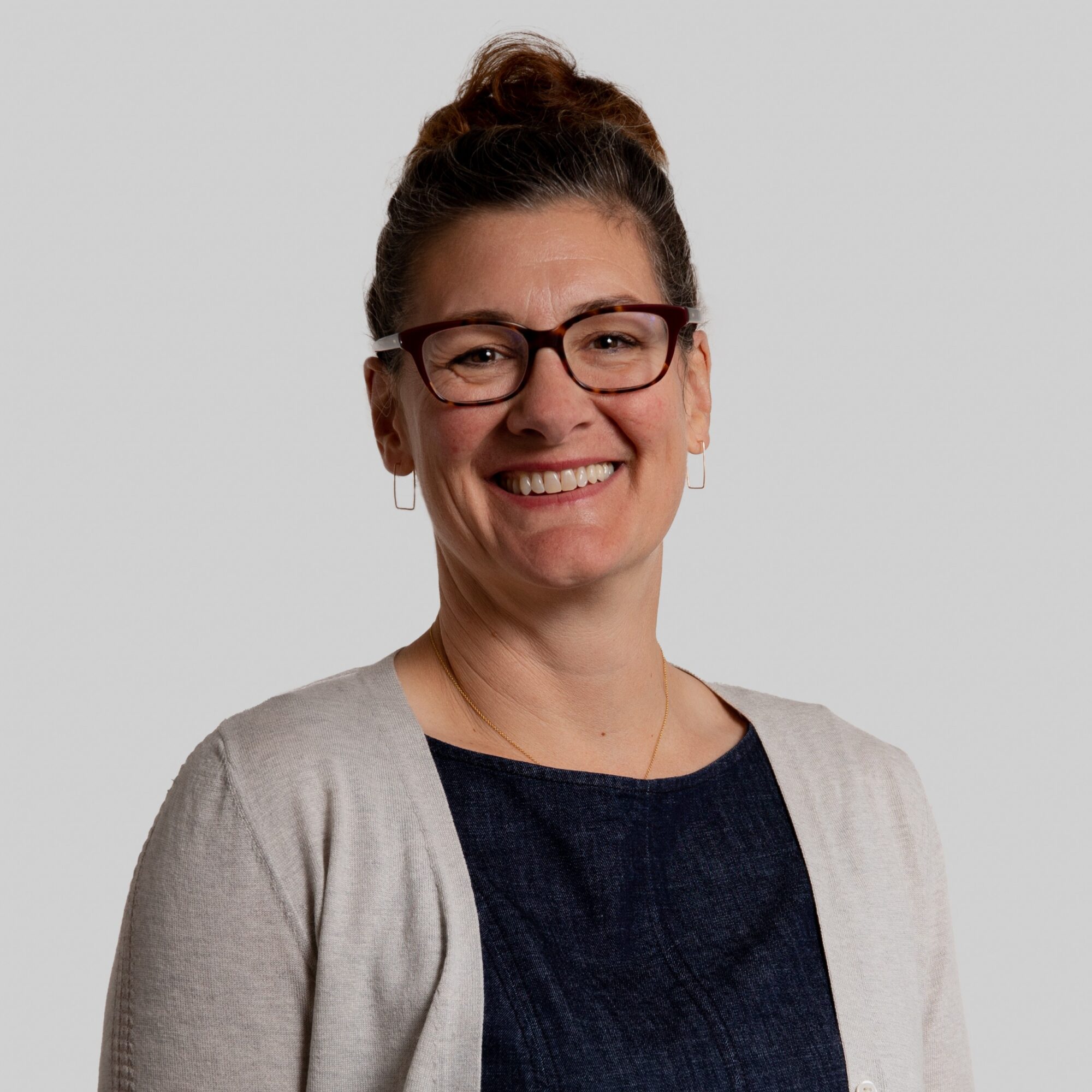 Smiling woman with glasses, wearing a light cardigan over a dark top, against a plain background.