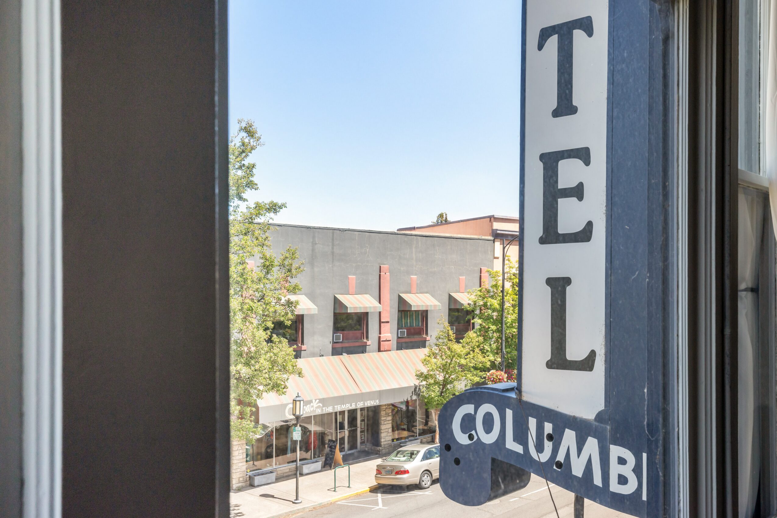 View through a window showing a street with buildings, trees, and parked cars, and a vertical sign reading 'TEL' and 'COLUMB'.