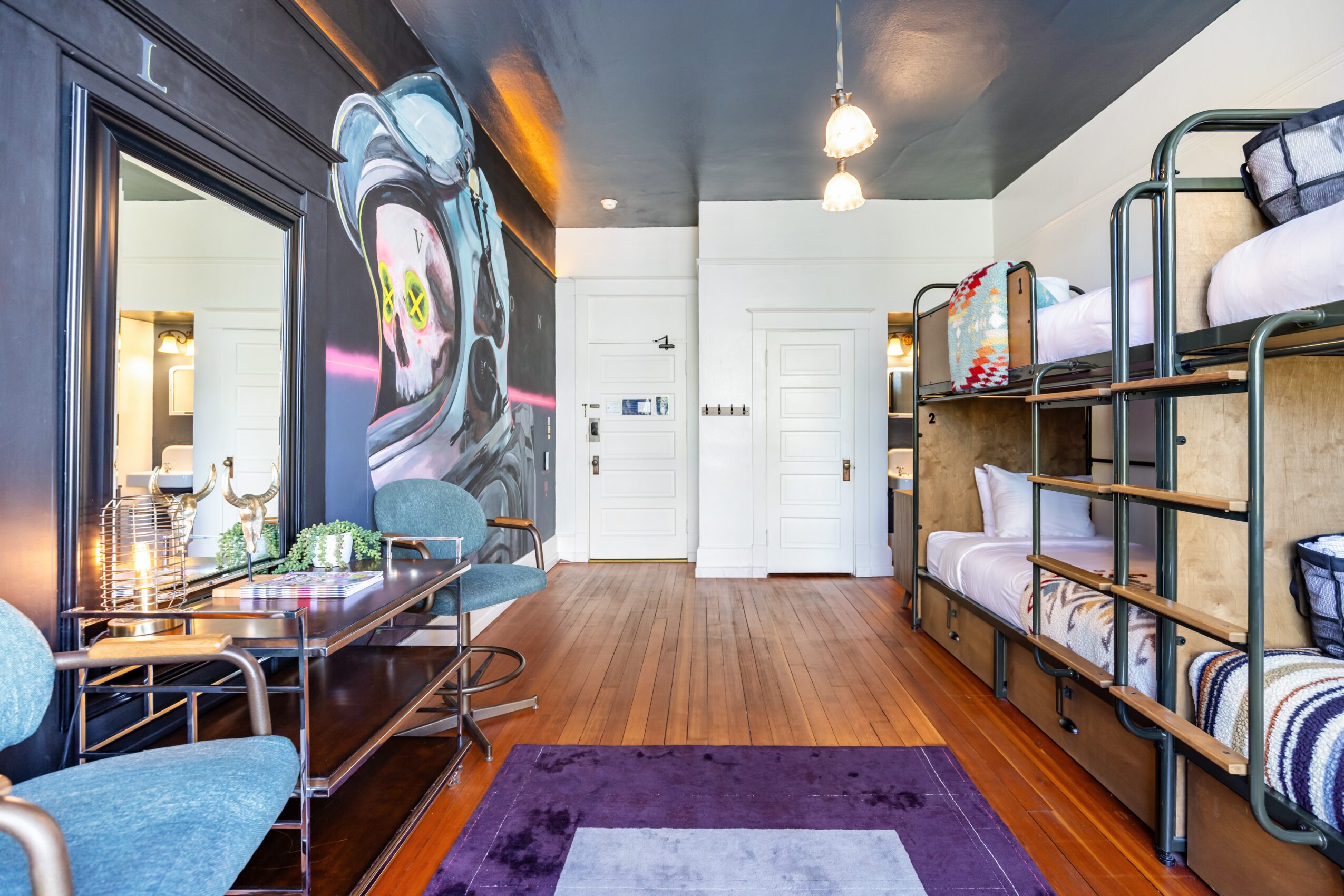 Room with a large graffiti mural, wooden floor, bunk beds, and a seating area with a table and blue chairs.