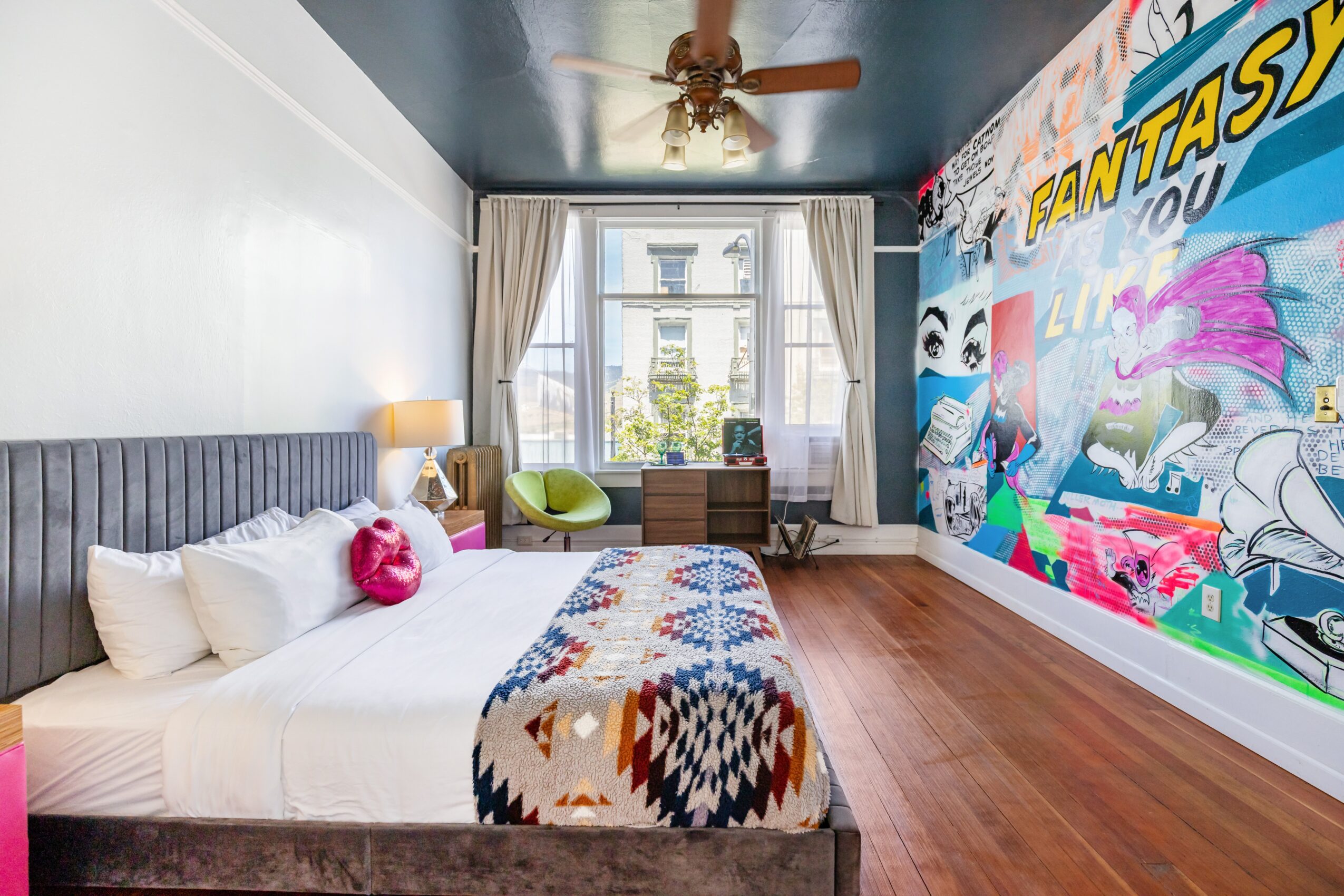 Bedroom with a bed, colorful wall mural, window with curtains, ceiling fan, and wooden floor.