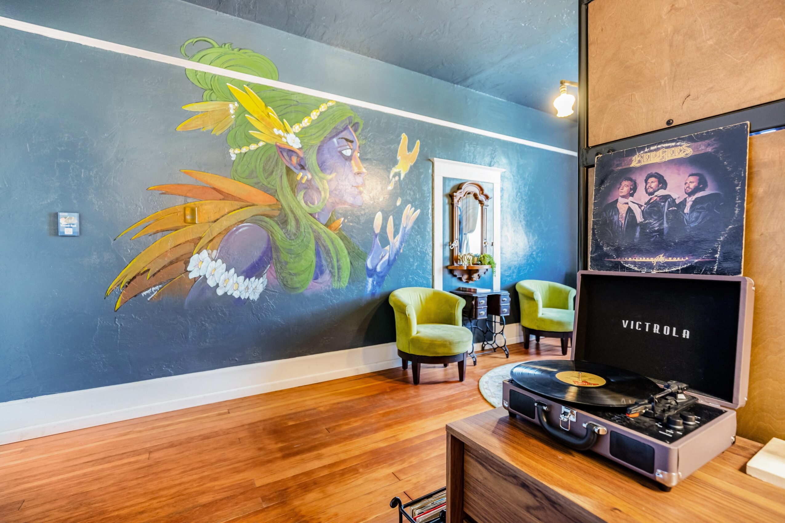 Room with colorful mural of a woman with feathers, two green chairs, a record player, and framed artwork on the wall.