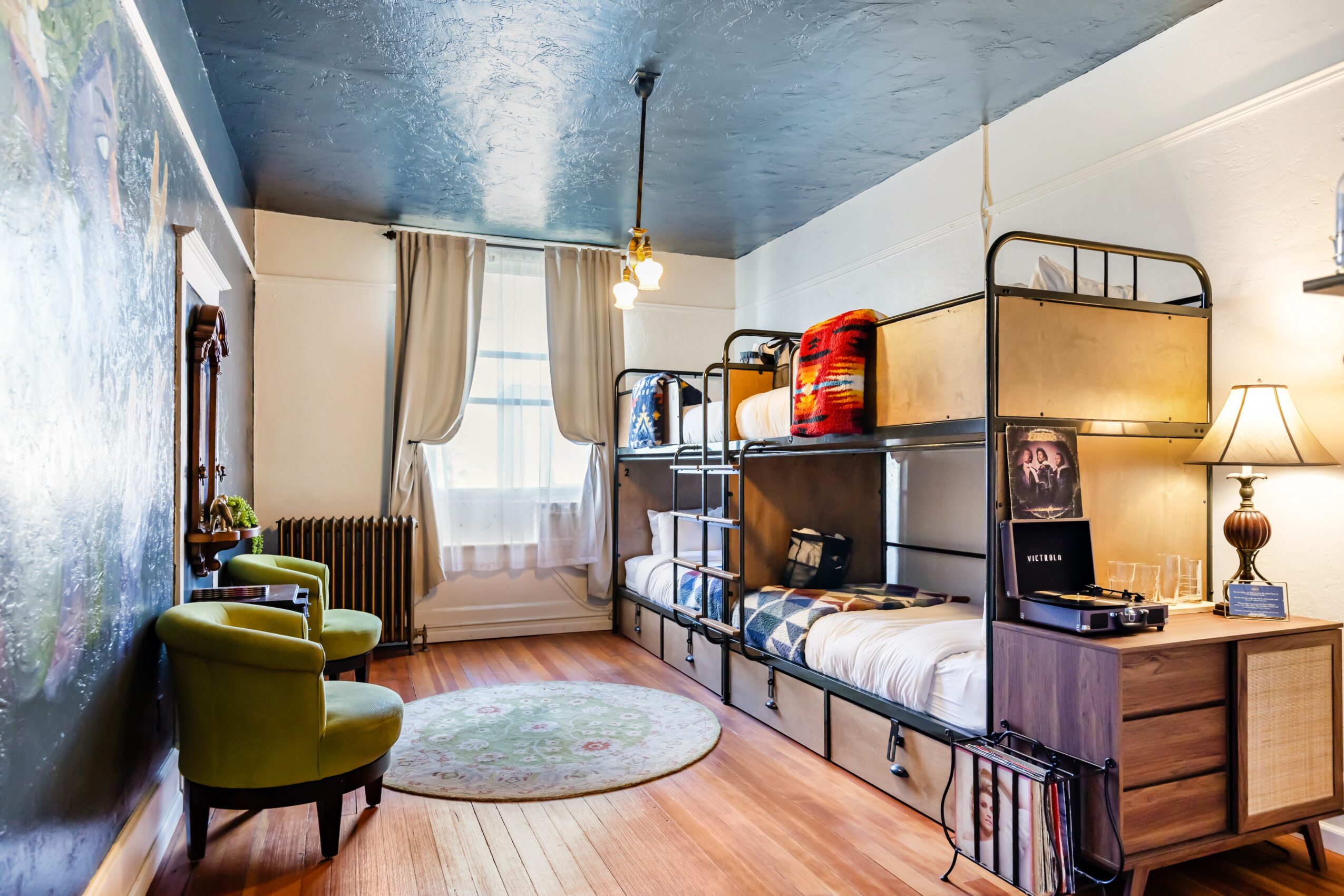 Room with bunk beds, green chairs, window with curtains, and a wooden floor.