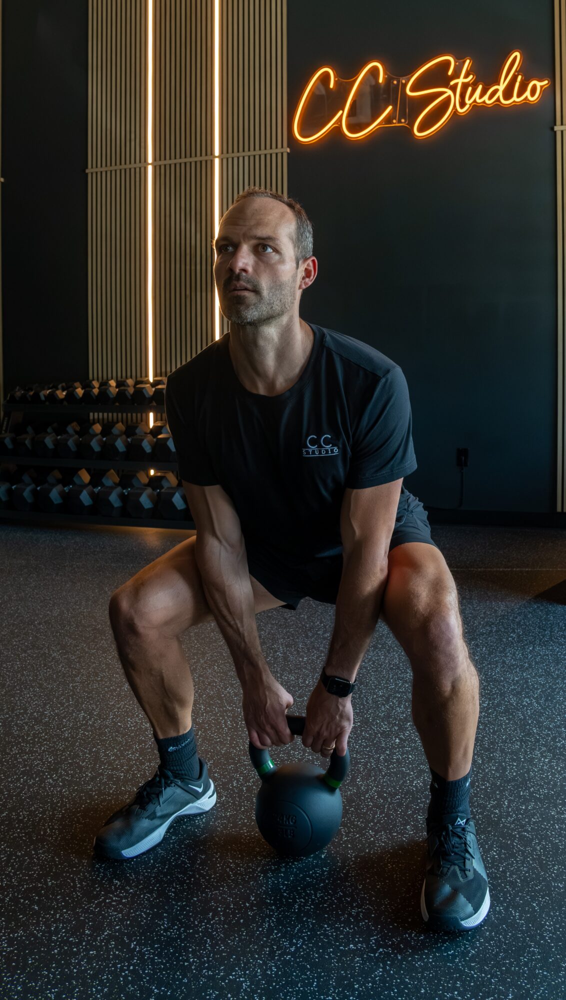 Man in gym holding kettlebell, neon sign reads 'CC Studio' in background.