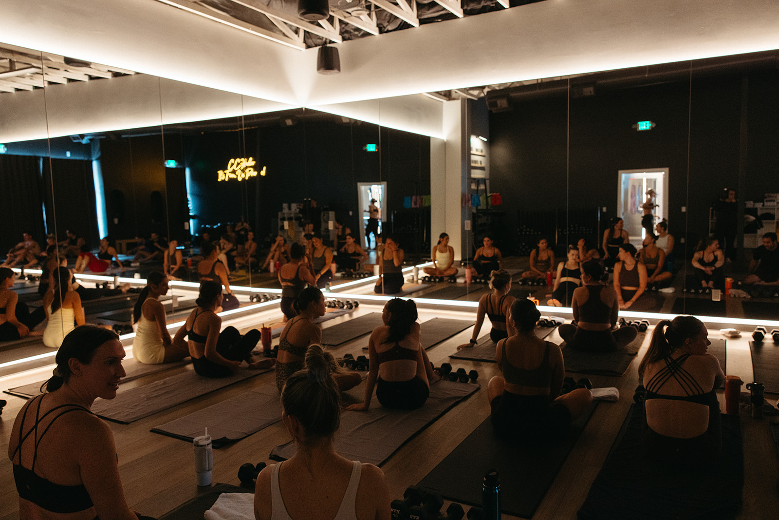 People participating in a yoga class in a dimly lit studio with mirrors and soft lighting, sitting on mats in a spacious room.