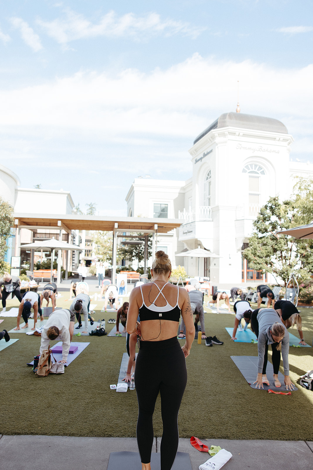 Group of people practicing yoga outdoors on mats in front of a white building, with a person in the foreground facing away.