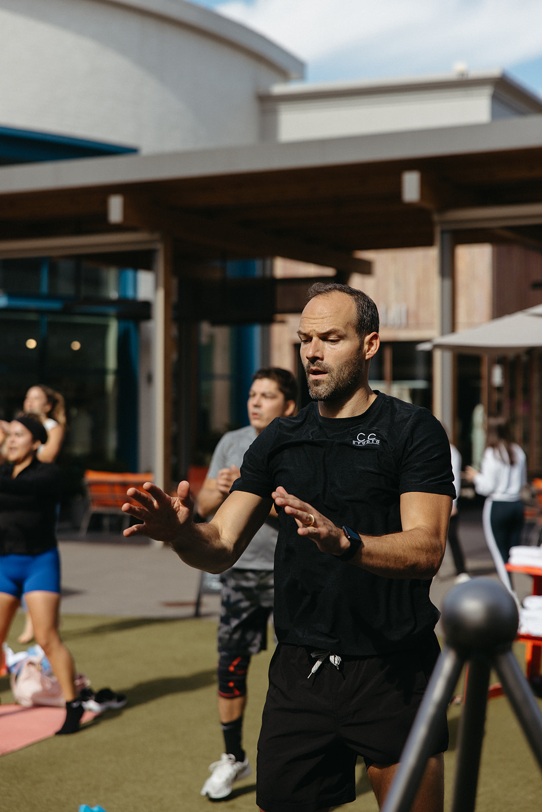 Man in black shirt and shorts gesturing outdoors with people in background under a building's overhang.