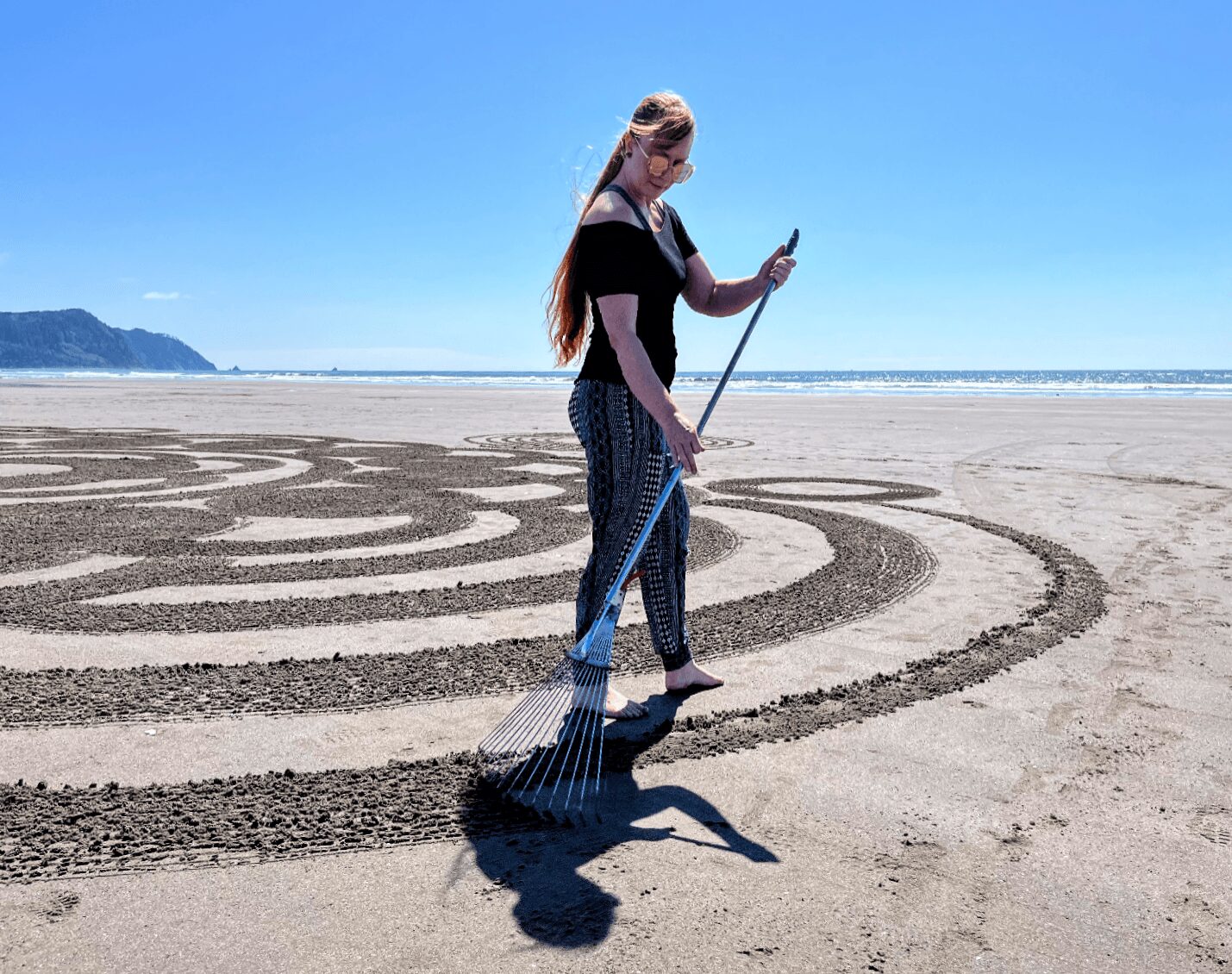 Woman standing on beach with a rake, looking at her phone, with sand maze patterns and ocean in background.