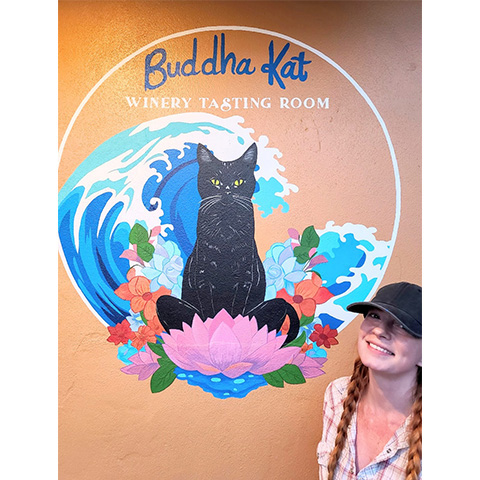 Person smiling in front of a colorful mural with a black cat, lotus flower, waves, and text 'Buddha Kat Winery Tasting Room'.