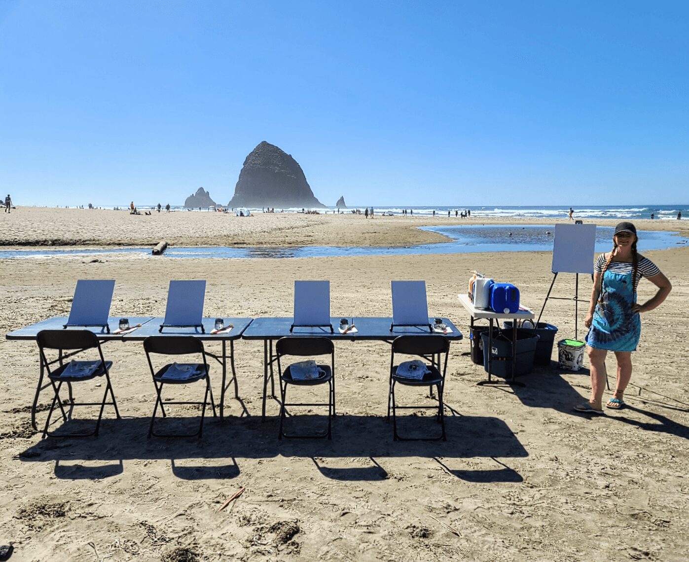 Beach scene with empty tables and chairs, a woman in a dress, and a large rock formation in the background.