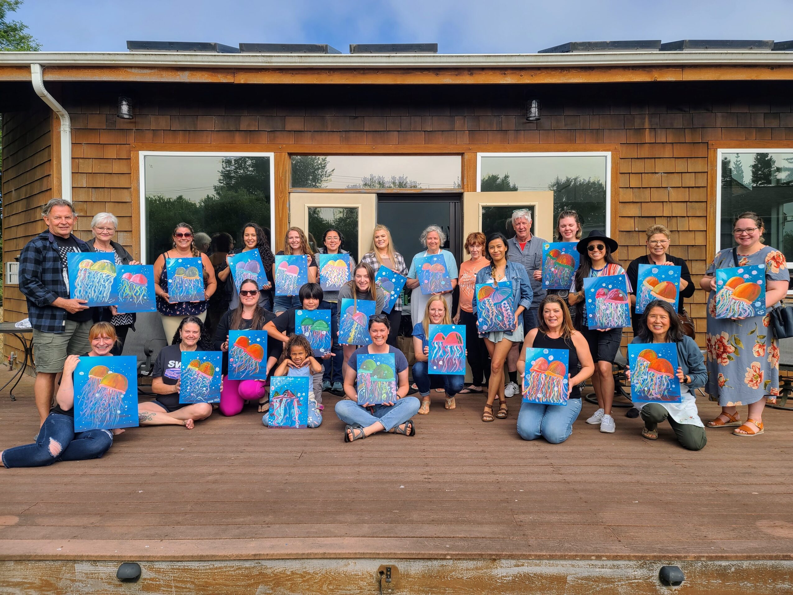 Group of people holding colorful paintings outdoors on a wooden deck in front of a building with large windows.