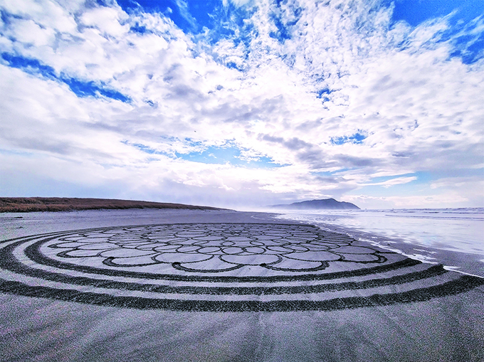 Sand art pattern on beach with sky and clouds above, distant land and water in background.