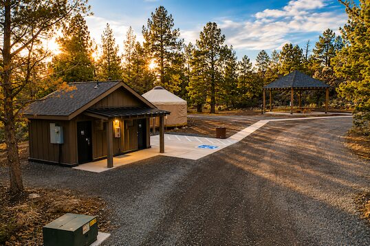 A small building with a dark roof and exterior, surrounded by trees and a paved parking area with a gazebo in the background.