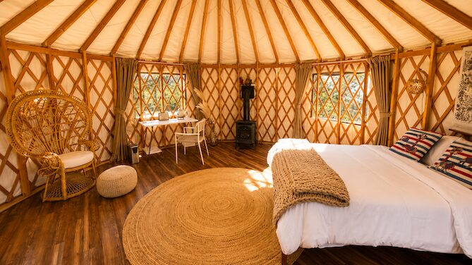 Interior of a yurt with a bed, wicker chair, round rug, and windows with curtains.