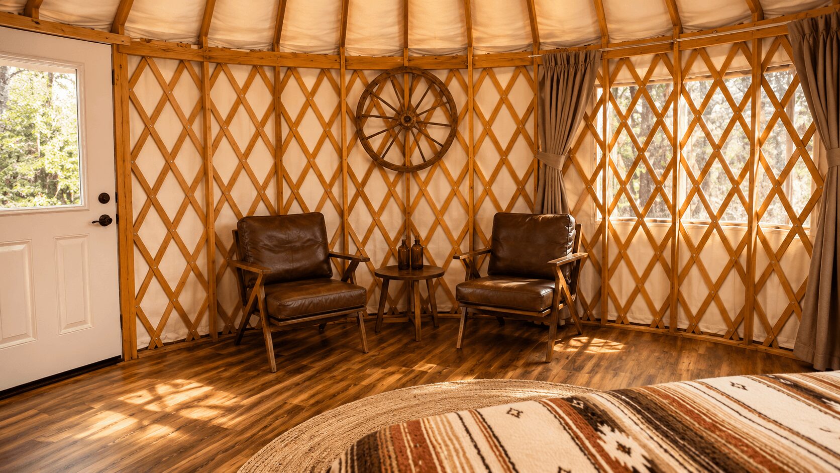 Interior of a yurt with two leather chairs, a small table, and a wheel decoration on the wall, with wooden flooring.