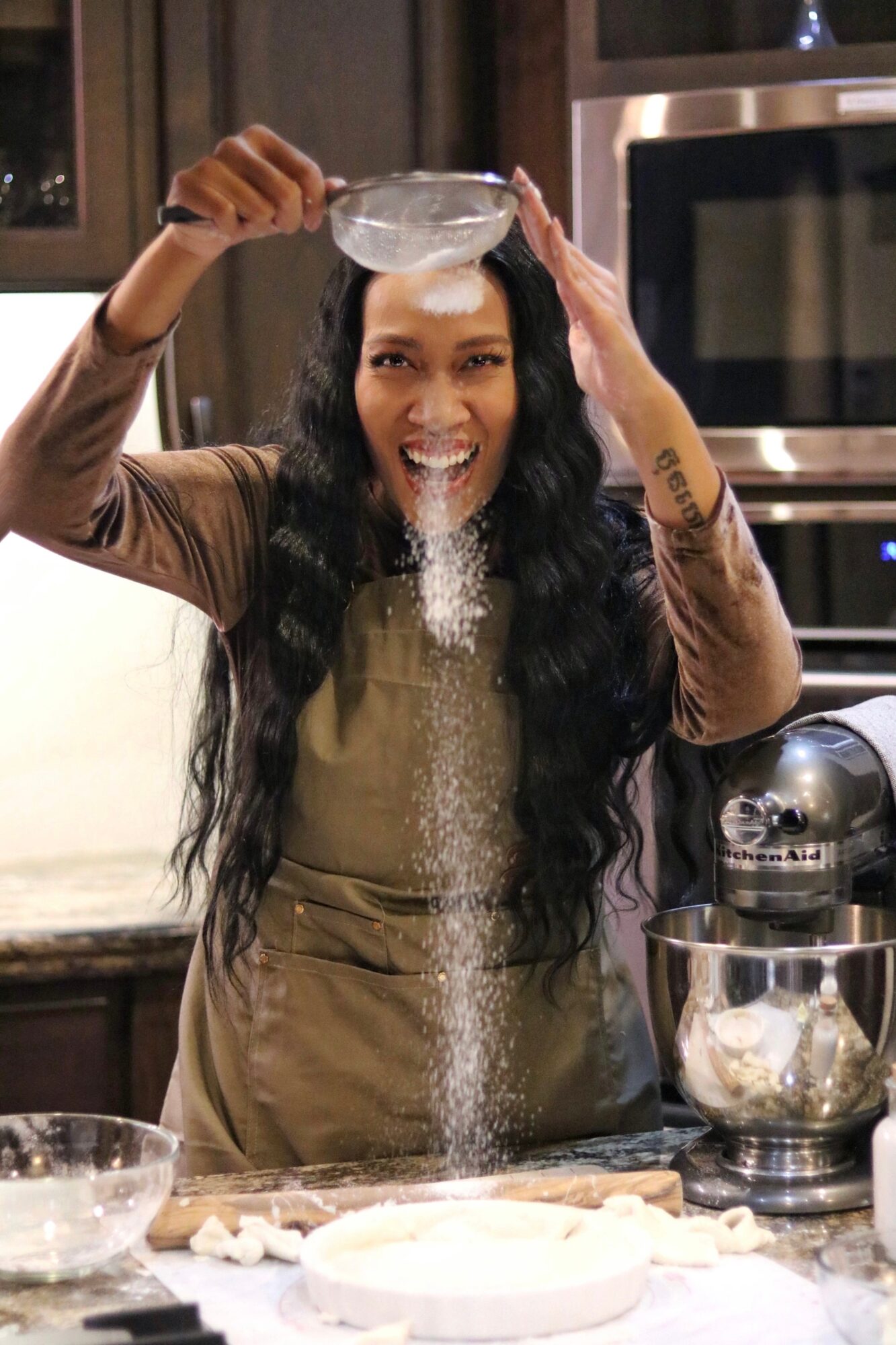 Woman in a kitchen pouring flour over dough, smiling, with baking tools around her.