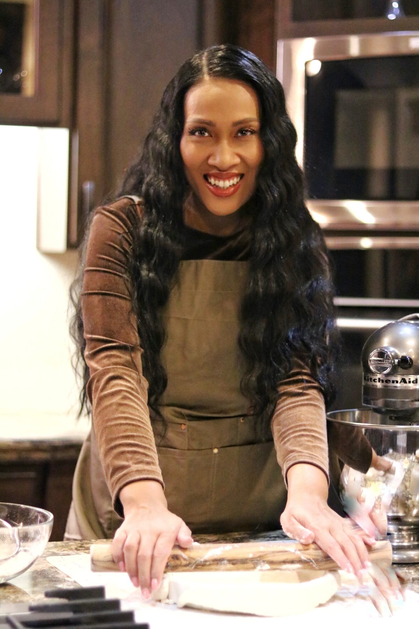 Woman with long wavy hair smiling in a kitchen, leaning on a counter with baking ingredients.