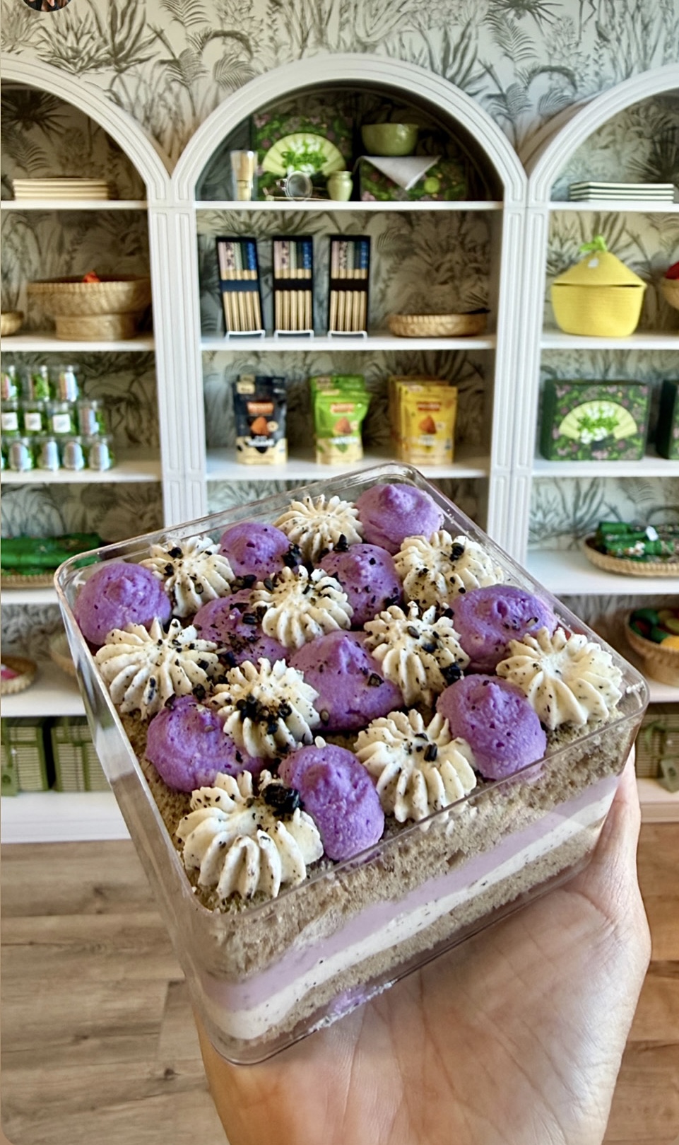 Hand holding a layered dessert with purple and white toppings, in front of a white cabinet with glass shelves.