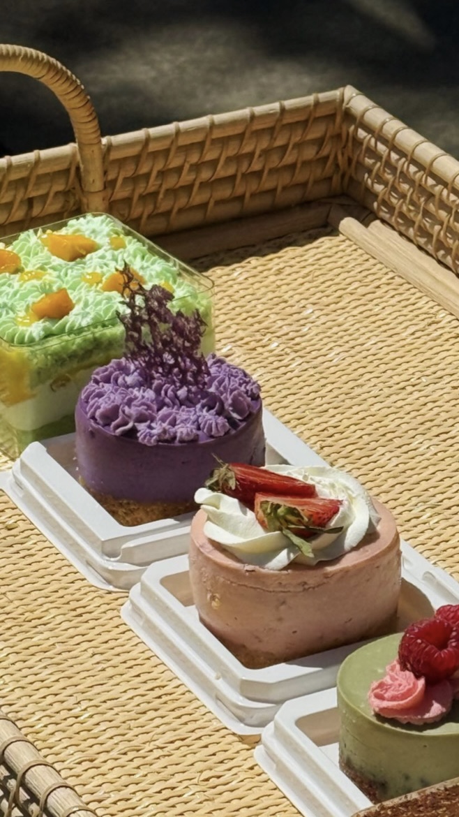 Four colorful cakes with various toppings on white plates in a woven basket, with a plain background.
