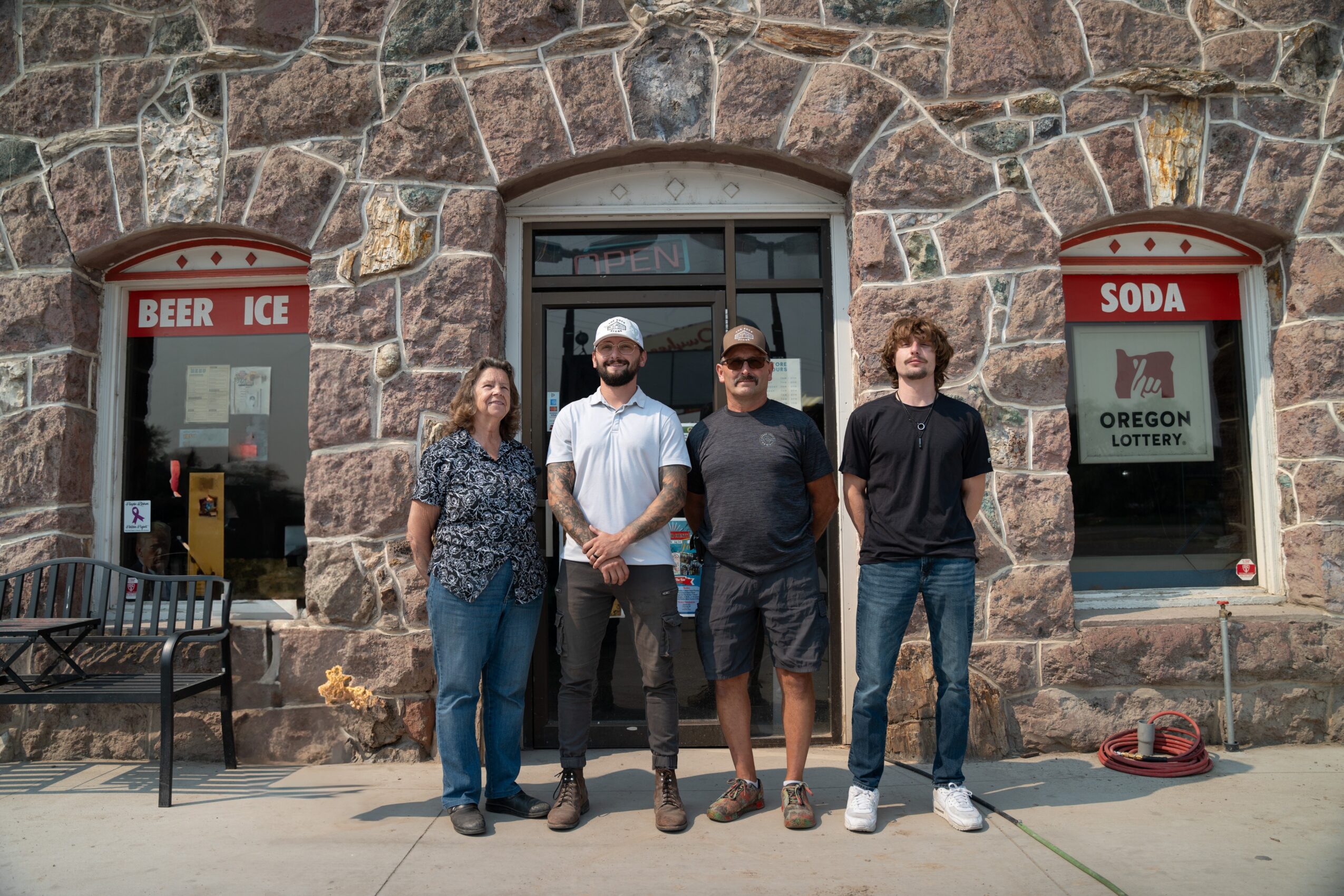 Four people stand in front of a stone building with two windows labeled 'BEER ICE' and 'SODA'.