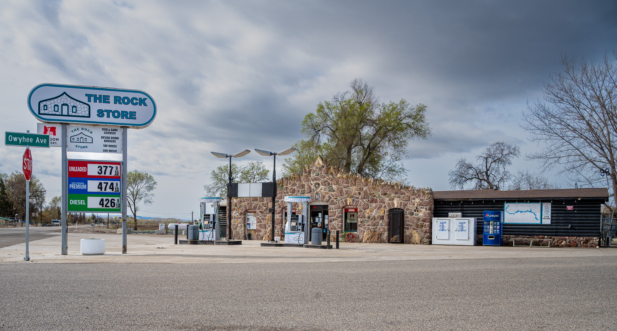 Gas station with brick building, trees, and cloudy sky, signs for fuel prices, and a road in foreground.