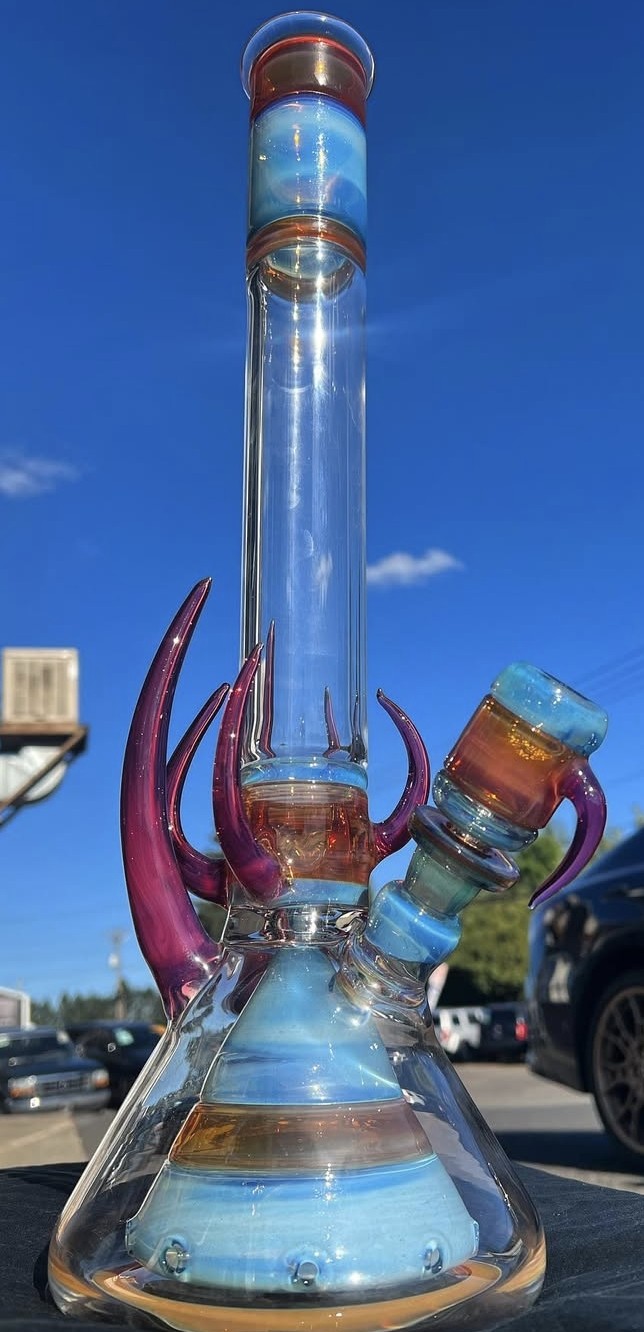 Glass bong with purple accents and a blue and orange bowl, outdoors with a parking lot and blue sky.