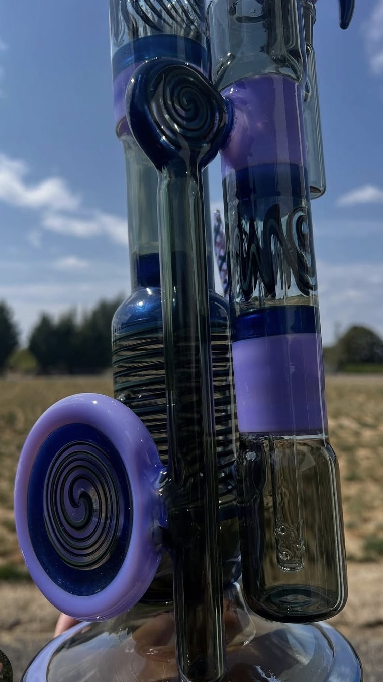 Close-up of glass pipes with purple and black spiral designs, outdoors with a blue sky and trees in the background.