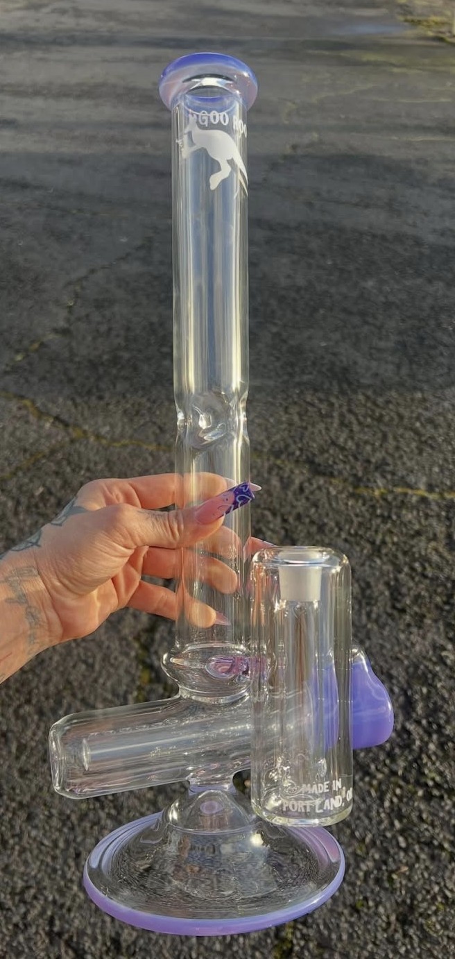 Person holding a glass bong outdoors on a paved surface, with a small glass container attached, purple accents visible.