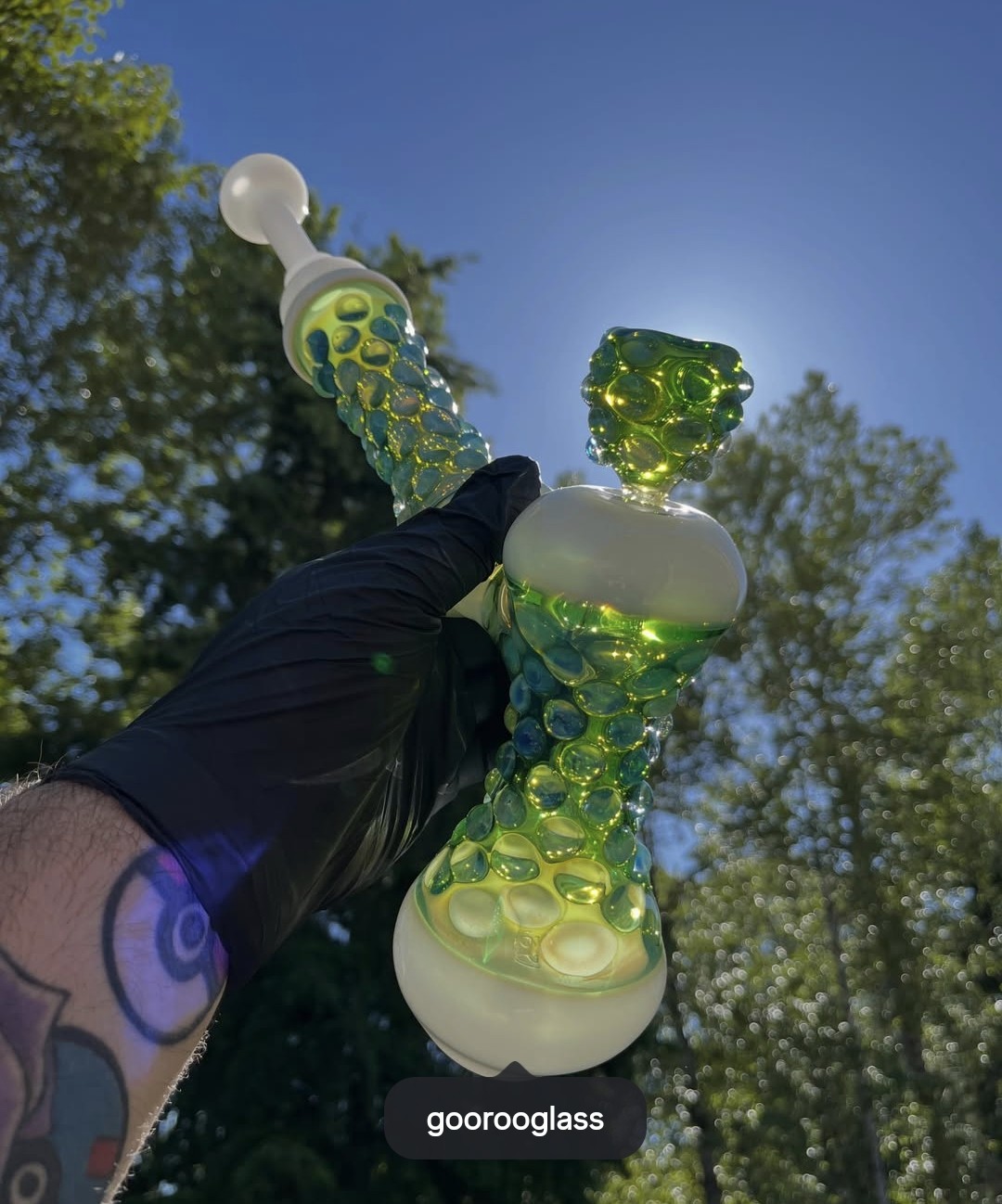 Person holding a colorful glass pipe outdoors with trees and blue sky in background.
