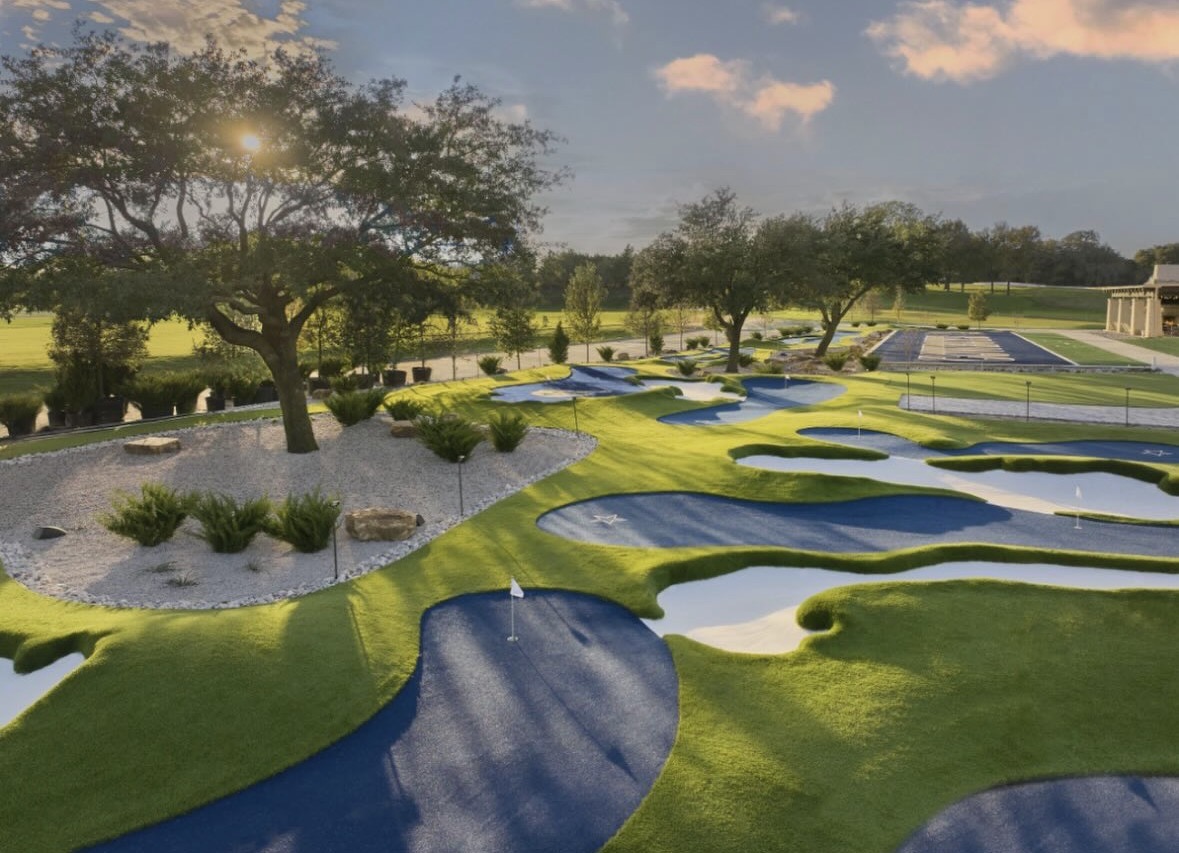 Miniature golf course with artificial water hazards, green grass, trees, and a building in the background under a partly cloudy sky.