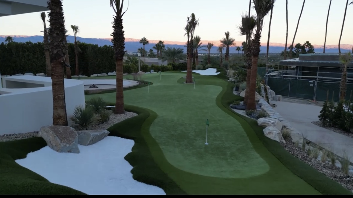 Golf course green with flag, surrounded by palm trees and landscaped areas, during sunset or dusk.