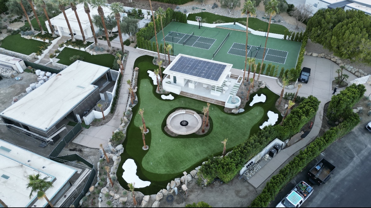 Aerial view of a property with tennis courts, a building, and landscaped yard with a circular feature, trees, and pathways.