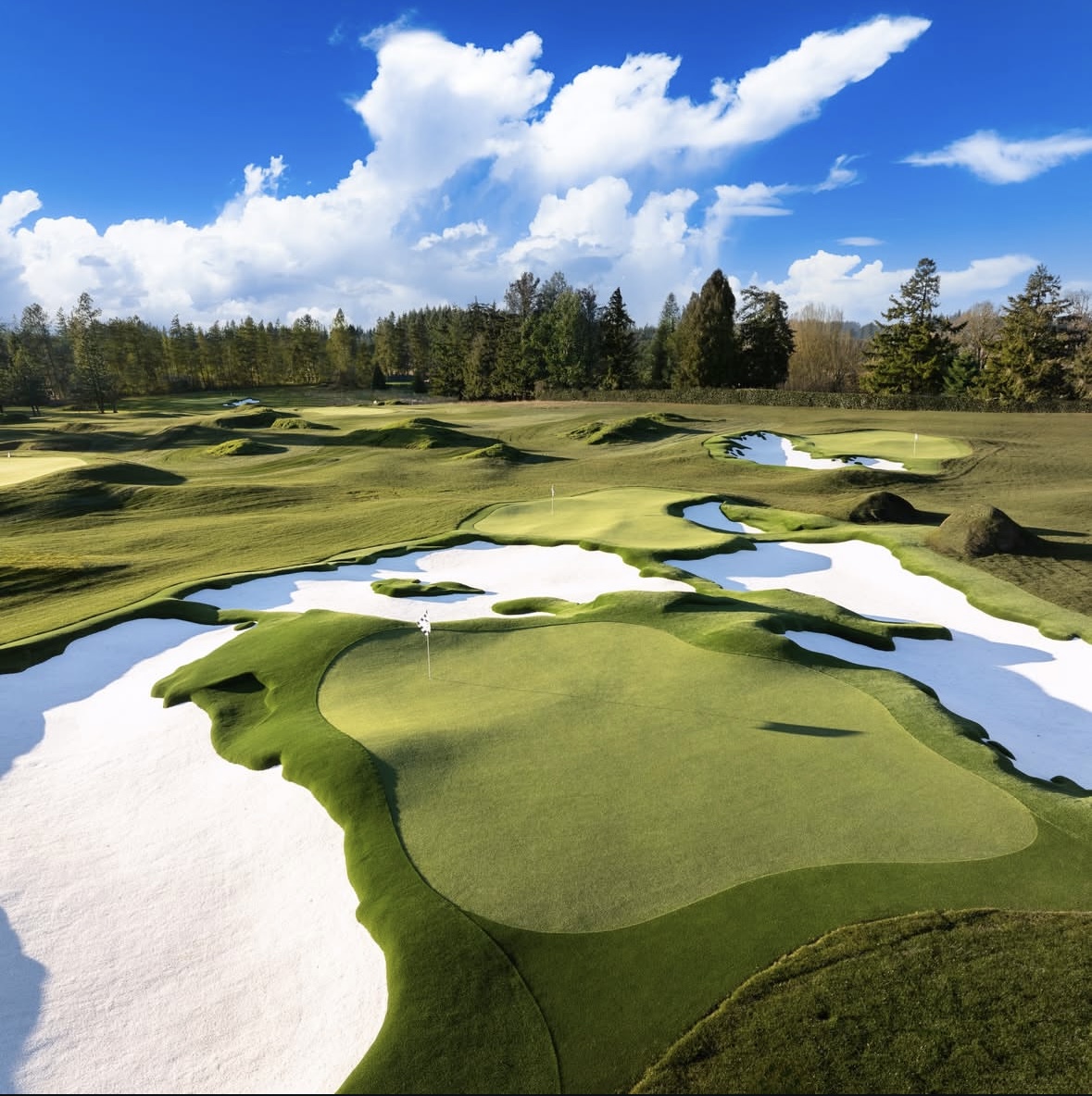Golf course with green fairways, sand traps, water hazards, and trees under a blue sky with clouds.