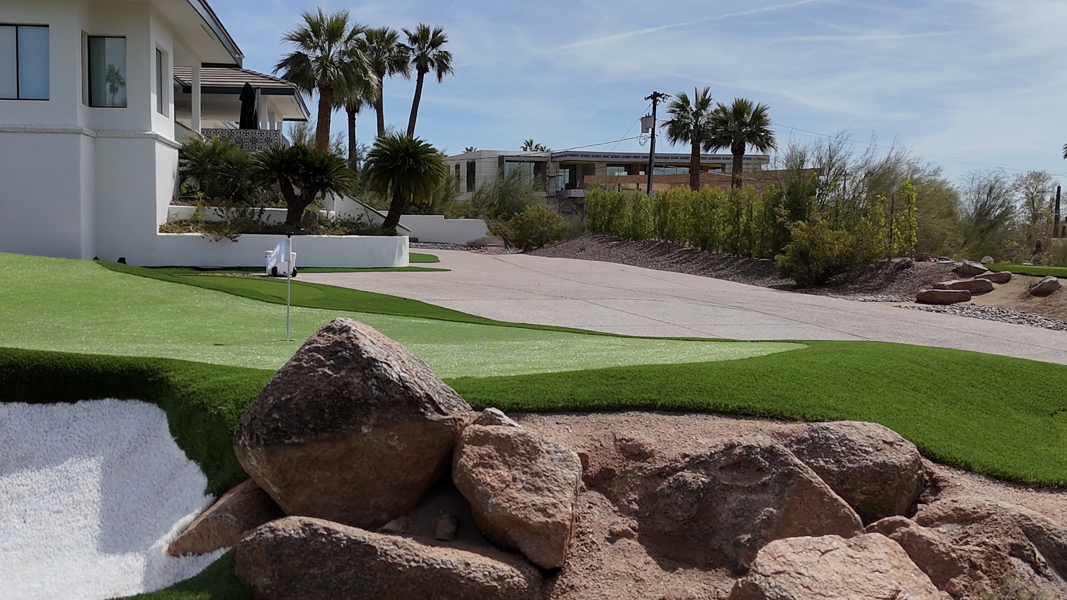Residential area with palm trees, a white house, and a landscaped yard with rocks and grass.