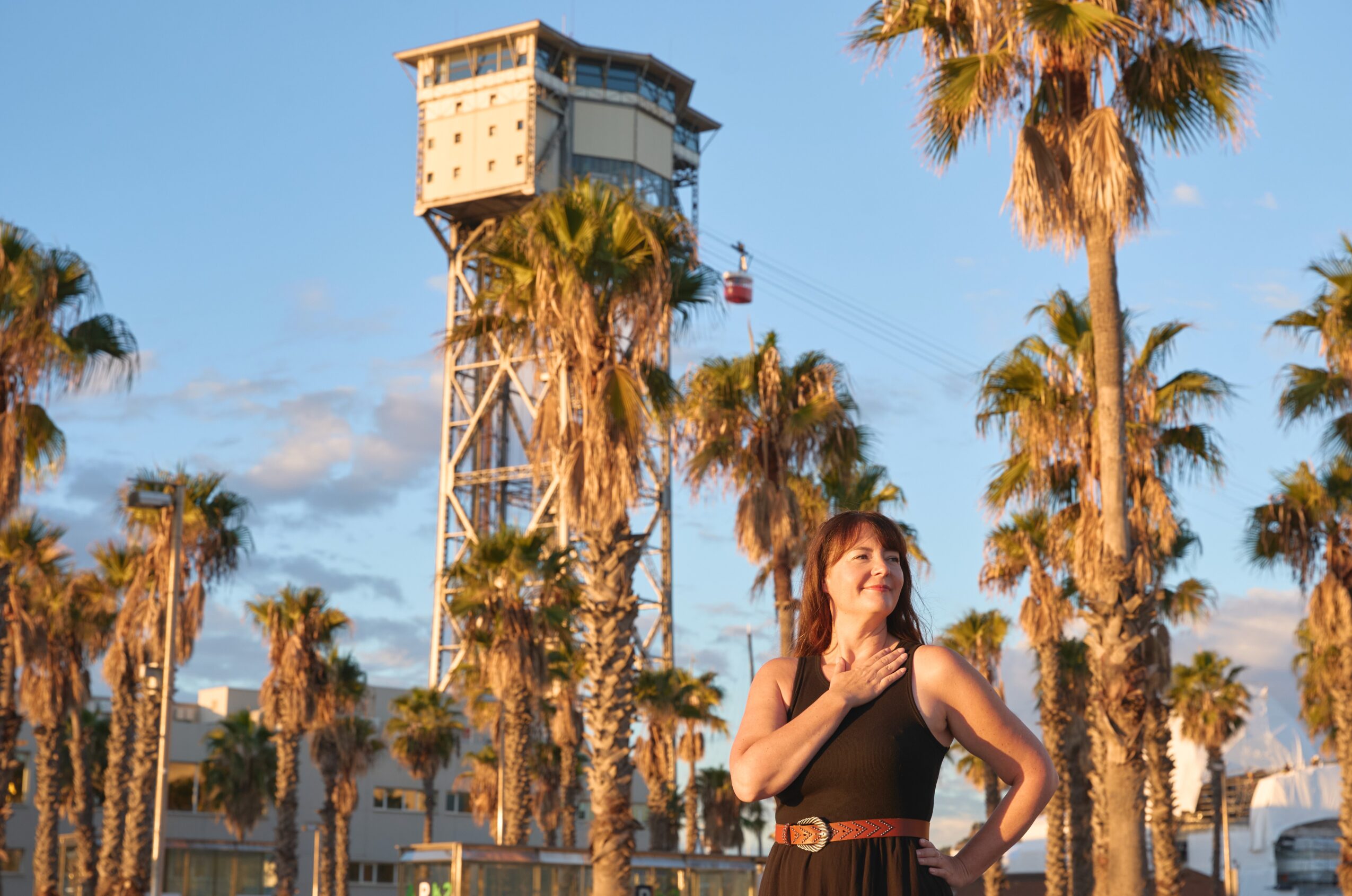 Woman standing among palm trees with a tower in the background, blue sky with clouds, sunny day.