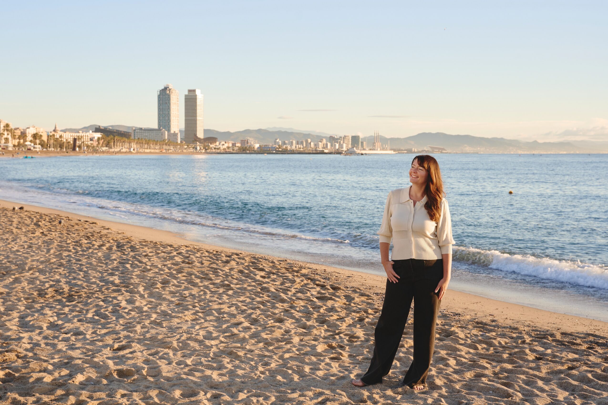 Woman standing on sandy beach near water with city skyline and mountains in background, sunny weather.