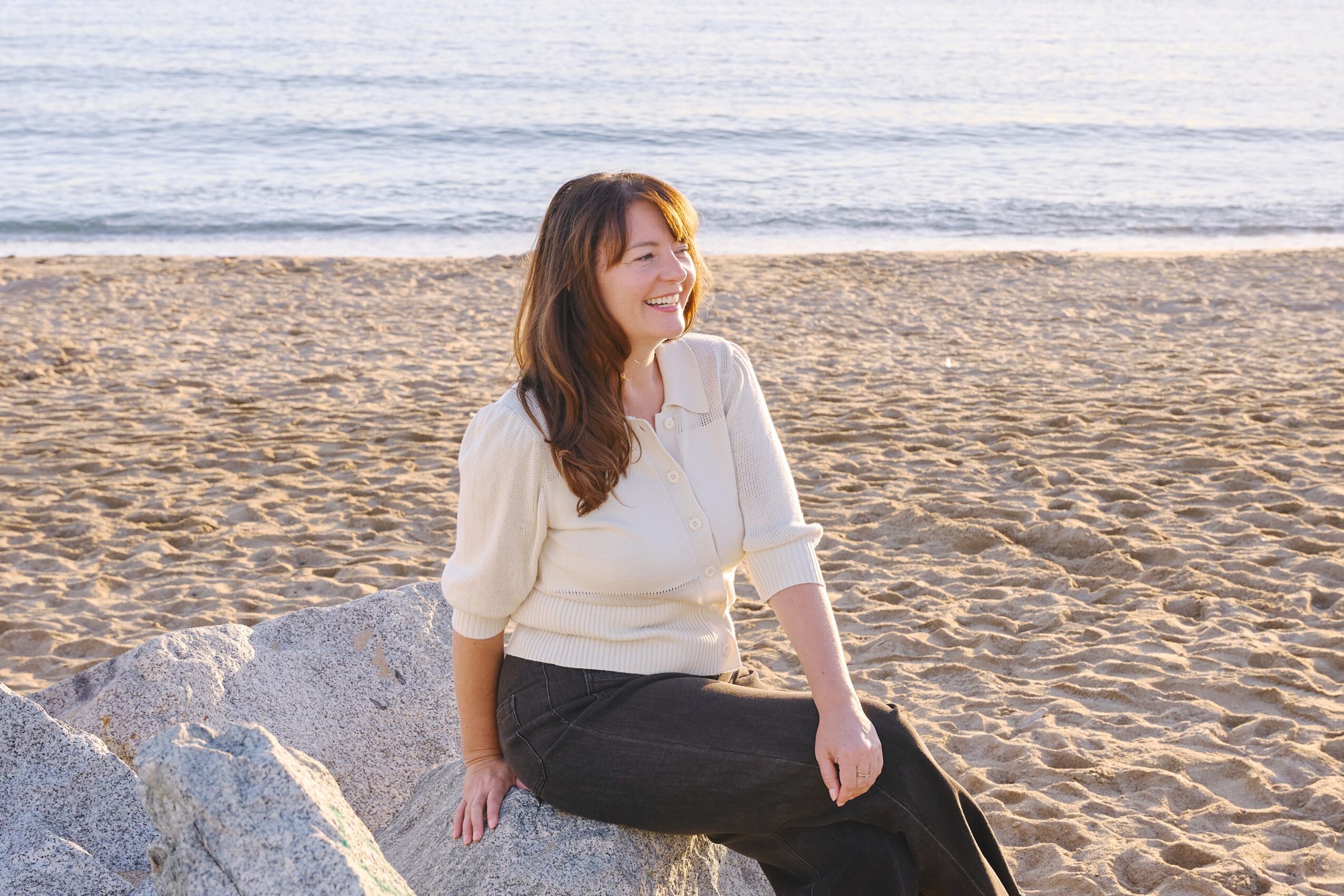 Woman sitting on a rock at the beach, smiling, with sand and water in the background.