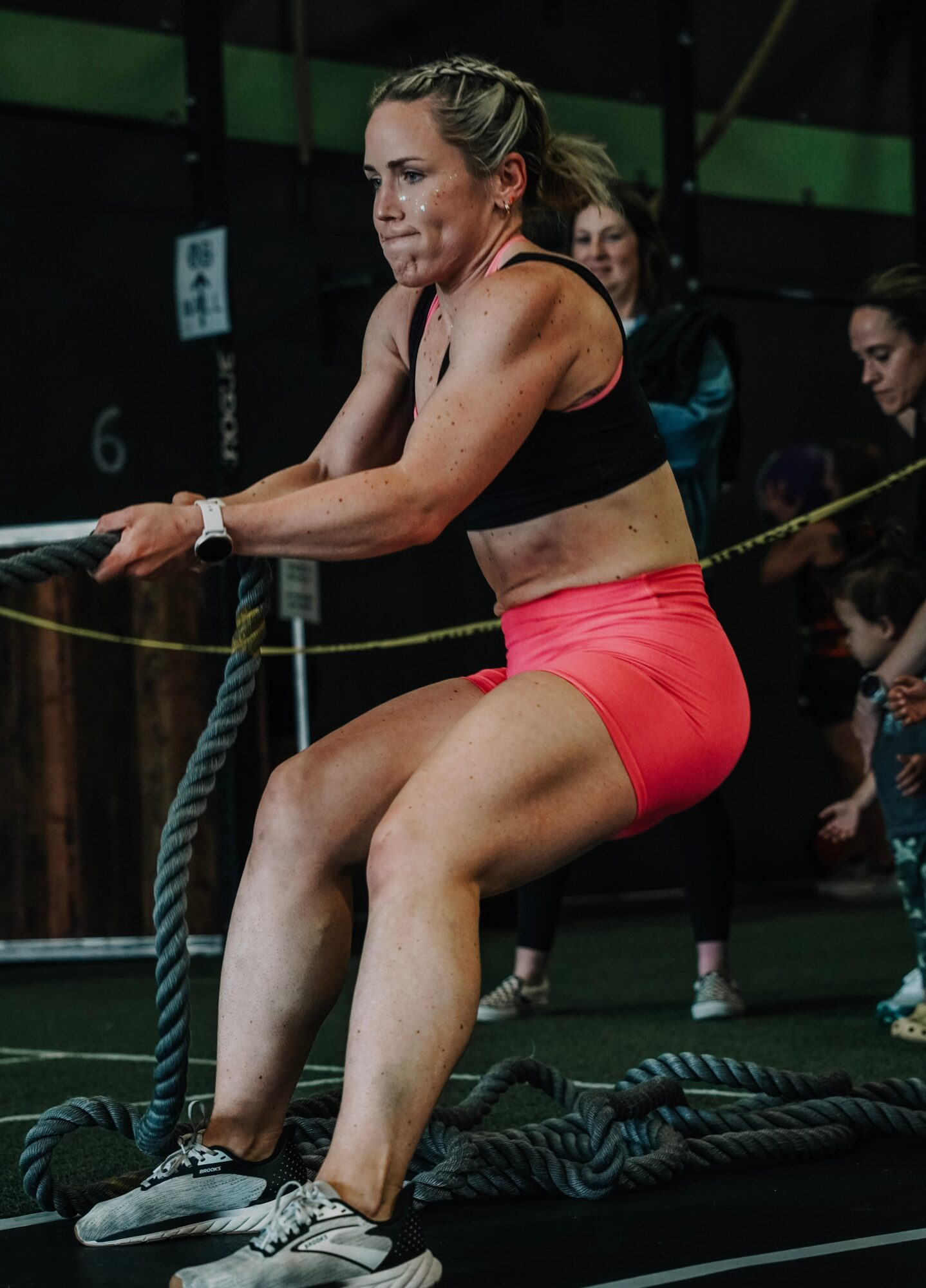 Woman in workout clothes pulling on thick ropes during exercise, with people in the background in a gym setting.