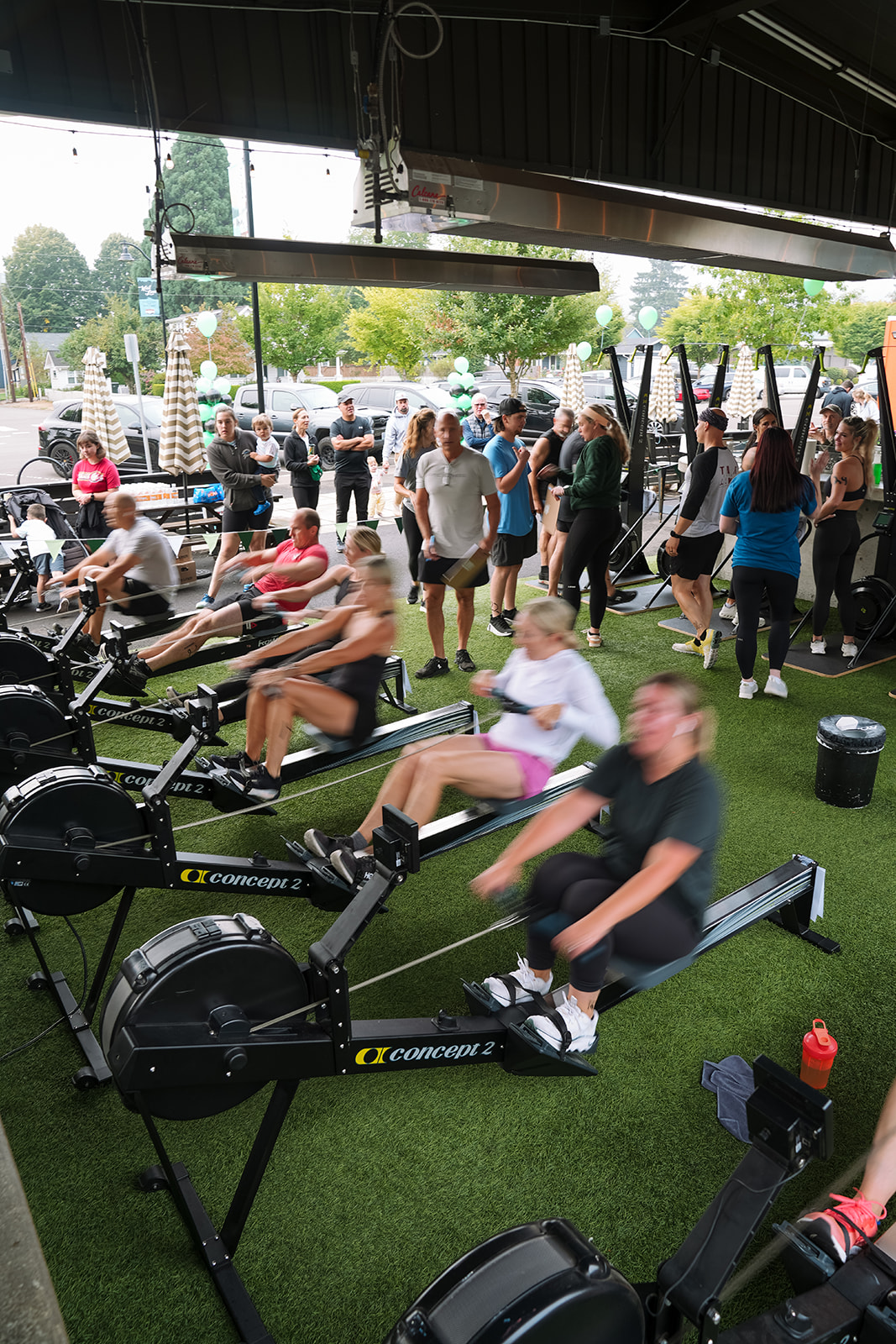 People exercising on rowing machines indoors, others standing and walking outside, trees and parked cars in background.